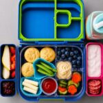Lunch du lundi dans une boîte à bento avec muffins salés, tortillons, pois mange-tout, concombres, poivrons orange, houmous et bleuets.